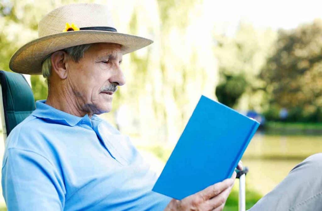 Ein älterer Mann mit Strohhut und hellblauem Hemd sitzt an einem sonnigen Tag im Freien und liest ein Buch mit blauem Einband, umgeben von viel Grün.