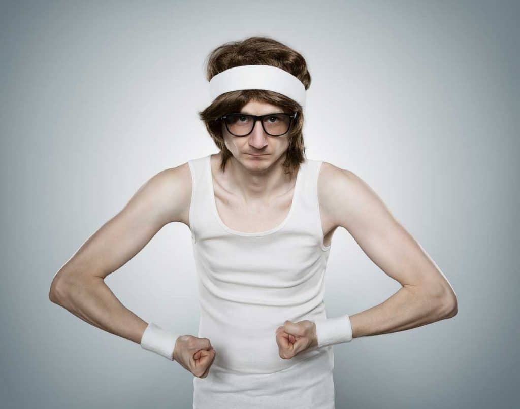 Eine schlanke Person, die ein weißes Tanktop, ein Stirnband, Schweißbänder und eine Brille trägt, posiert mit angespannten Armen und ernstem Gesichtsausdruck vor einem schlichten Hintergrund.