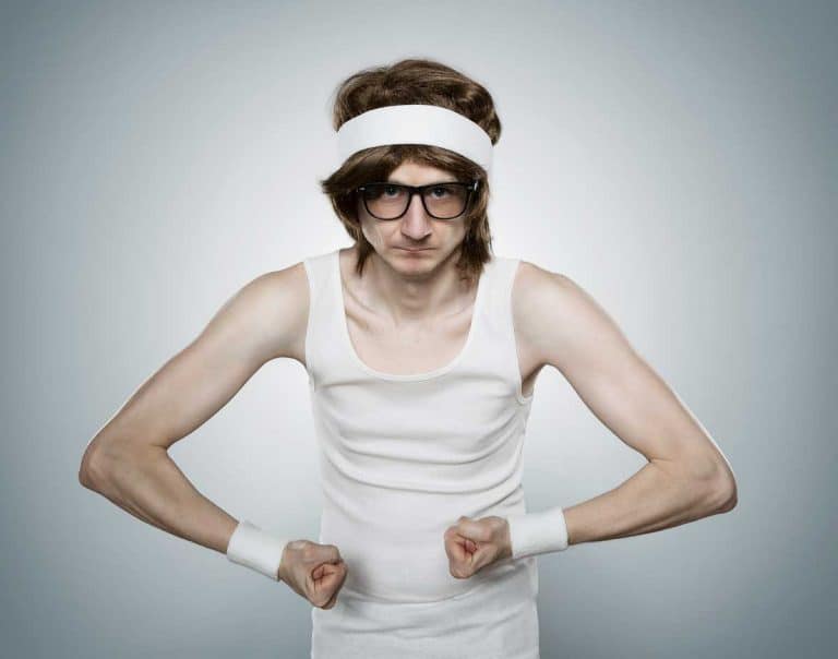 Eine schlanke Person, die ein weißes Tanktop, ein Stirnband, Schweißbänder und eine Brille trägt, posiert mit angespannten Armen und ernstem Gesichtsausdruck vor einem schlichten Hintergrund.