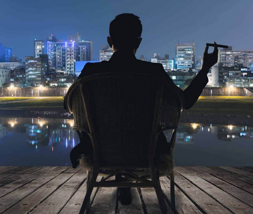 Eine Person sitzt nachts auf einem Stuhl auf einer Holzterrasse, hält eine Zigarre in der Hand und blickt über einen Fluss hinweg auf die Skyline der Stadt, die sich als Silhouette vor den beleuchteten Gebäuden abzeichnet.