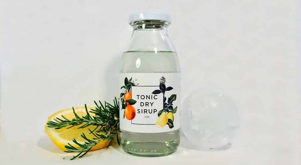 Eine durchsichtige Glasflasche mit der Aufschrift „Tonic Dry Sirup“ und Abbildungen von Zitrusfrüchten, einer Zitronenscheibe, einem Rosmarinzweig und einem runden Eiswürfel auf weißem Hintergrund.