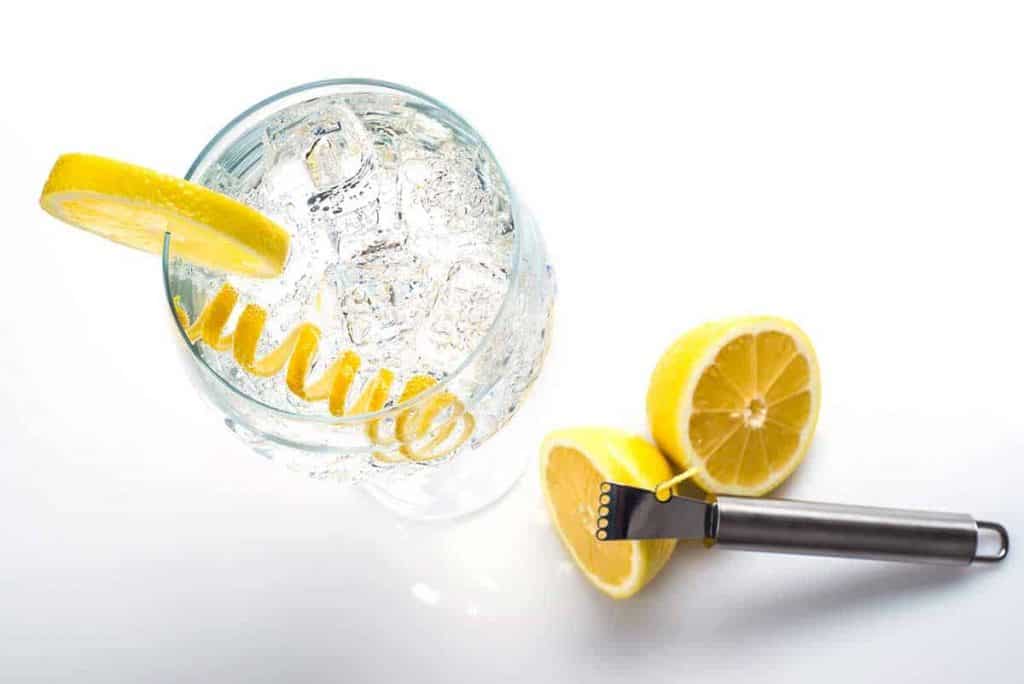 Ein Glas Sprudelwasser mit Eis, einer Zitronenscheibe und einer Zitronenzeste. Neben dem Glas liegen zwei Zitronenhälften und ein Zitronenreibe aus Metall vor einem weißen Hintergrund.