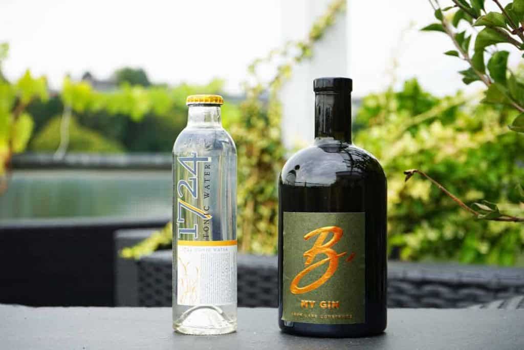 Eine Flasche „1724 Tonic Water“ und eine Flasche „B My Gin“ stehen nebeneinander auf einem Tisch im Freien; im unscharfen Hintergrund sind Grünpflanzen und Wasser zu sehen.