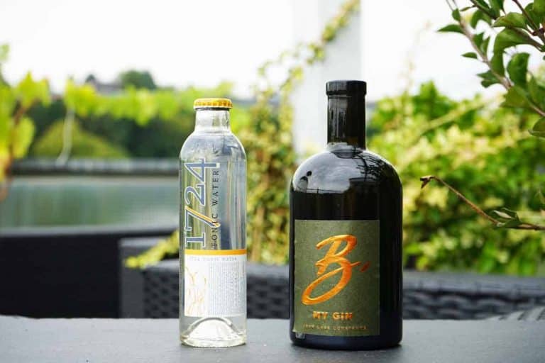 Eine Flasche „1724 Tonic Water“ und eine Flasche „B My Gin“ stehen nebeneinander auf einem Tisch im Freien; im unscharfen Hintergrund sind Grünpflanzen und Wasser zu sehen.