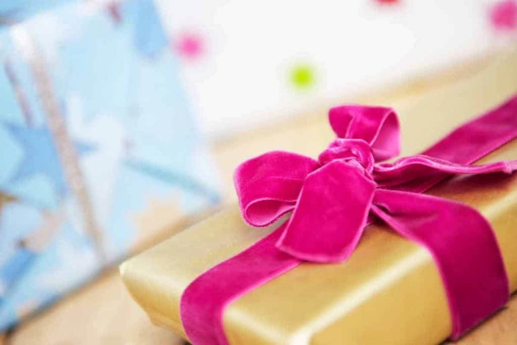 Eine in Goldpapier eingepackte Geschenkbox mit einer leuchtend rosa Schleife aus Samt. Im Hintergrund ist ein weiteres, unscharfes Geschenk in blauer Verpackung zu sehen.