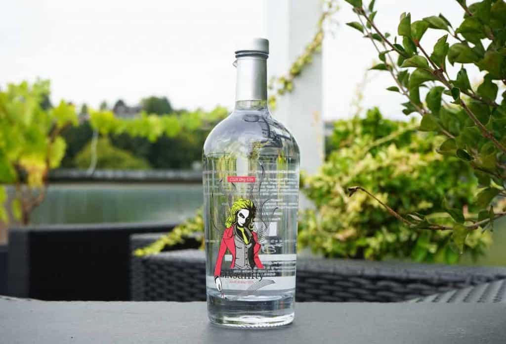 Eine durchsichtige Gin-Flasche mit einem Etikett, auf dem eine Frau in einer roten Jacke abgebildet ist, steht auf einem Tisch im Freien, im Hintergrund sind Pflanzen und Wasser zu sehen.