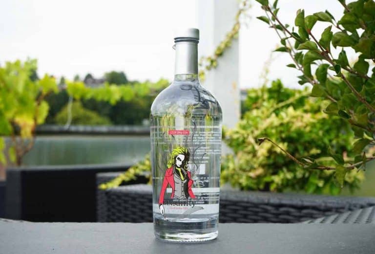 Eine durchsichtige Gin-Flasche mit einem Etikett, auf dem eine Frau in einer roten Jacke abgebildet ist, steht auf einem Tisch im Freien, im Hintergrund sind Pflanzen und Wasser zu sehen.