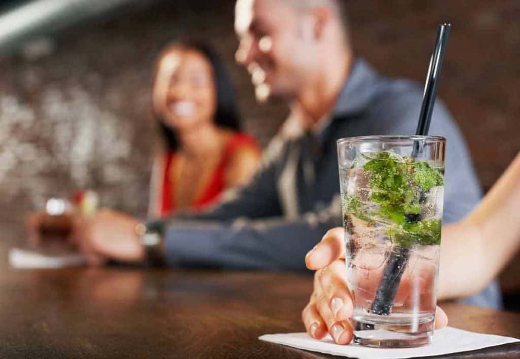 Eine Nahaufnahme einer Hand, die einen Mojito-Cocktail auf einer Serviette in einer Bar hält, während im unscharfen Hintergrund ein lächelnder Mann und eine lächelnde Frau nebeneinander sitzen.
