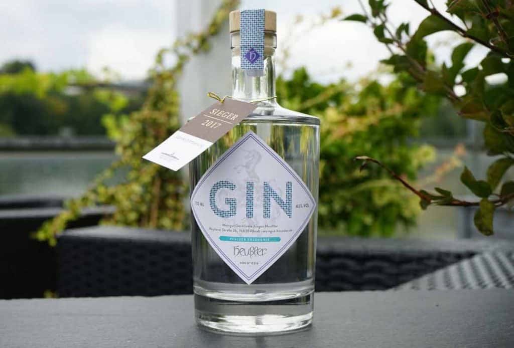 Auf einem Tisch im Freien steht eine Gin-Flasche aus klarem Glas mit einem weißen Etikett, umgeben von grünen Pflanzen, während im Hintergrund ein Gewässer und Bäume verschwommen zu sehen sind.
