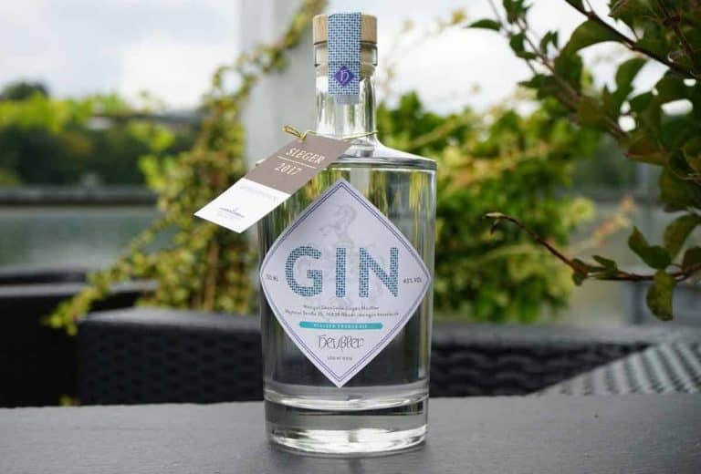 Auf einem Tisch im Freien steht eine Gin-Flasche aus klarem Glas mit einem weißen Etikett, umgeben von grünen Pflanzen, während im Hintergrund ein Gewässer und Bäume verschwommen zu sehen sind.