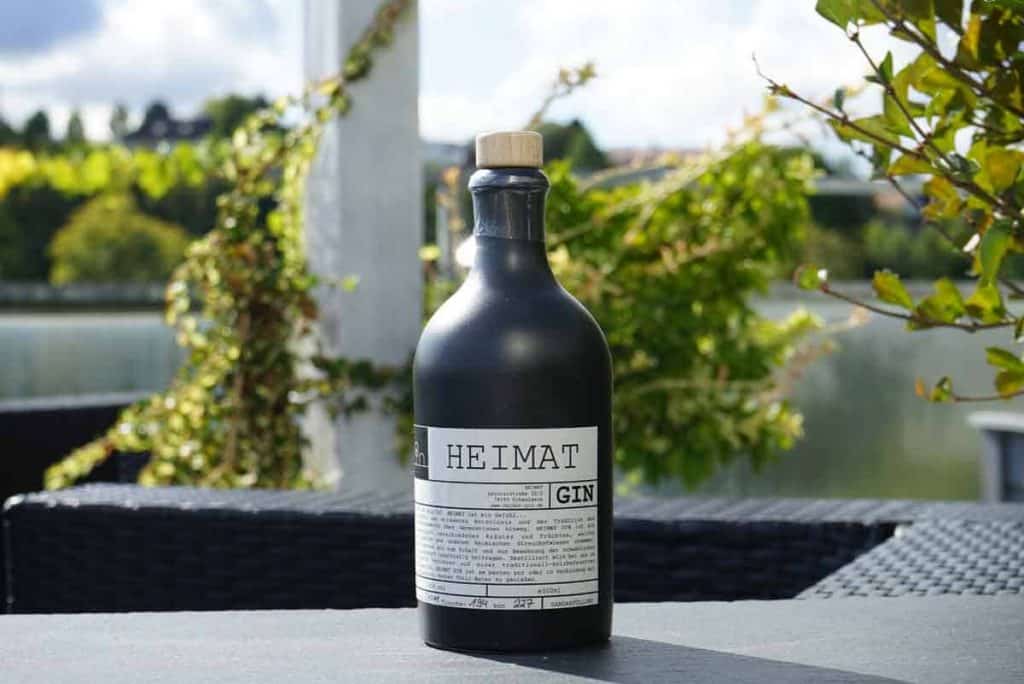Auf einem dunklen Tisch im Freien steht eine schwarze Flasche mit der Aufschrift „HEIMAT GIN“, im Hintergrund sind Grünflächen, ein See und ein teilweise bewölkter Himmel zu sehen.