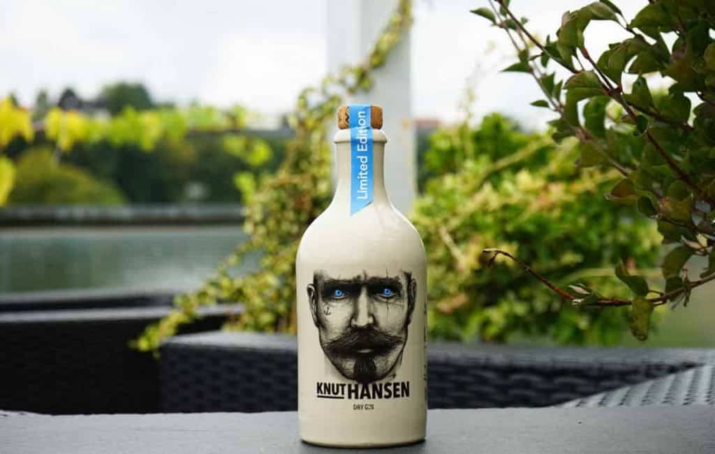 Eine Flasche „Knut Hansen Dry Gin“ in limitierter Auflage, auf deren Etikett das Gesicht eines bärtigen Mannes abgebildet ist, steht auf einem Tisch im Freien, im Hintergrund sind grüne Pflanzen und Wasser zu sehen.