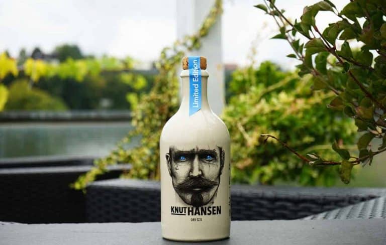 Eine Flasche „Knut Hansen Dry Gin“ in limitierter Auflage, auf deren Etikett das Gesicht eines bärtigen Mannes abgebildet ist, steht auf einem Tisch im Freien, im Hintergrund sind grüne Pflanzen und Wasser zu sehen.