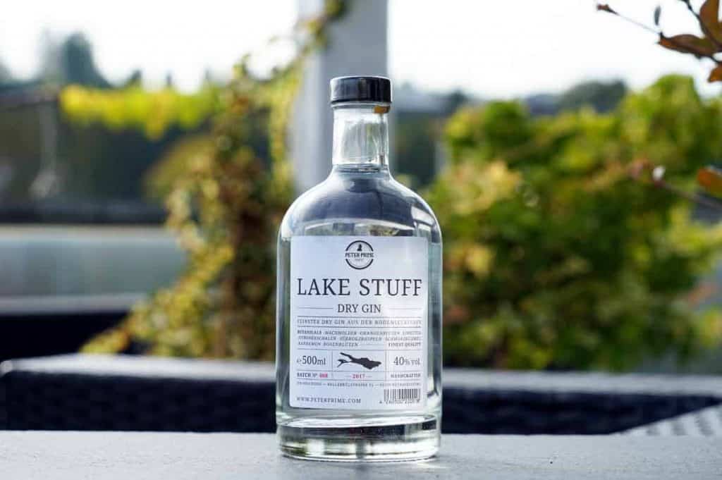 Auf einem dunklen Tisch im Freien steht eine durchsichtige Flasche „Lake Stuff Dry Gin“, im Hintergrund sind Grünpflanzen und verschwommene Blätter zu sehen. Auf dem Etikett sind die Markennamen und der Alkoholgehalt angegeben.
