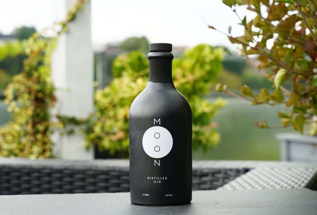 Auf einem Tisch im Freien steht eine schwarze Flasche mit der Aufschrift „MOON DISTILLED GIN“, im Hintergrund sind grüne Pflanzen und verschwommene Bäume zu sehen.