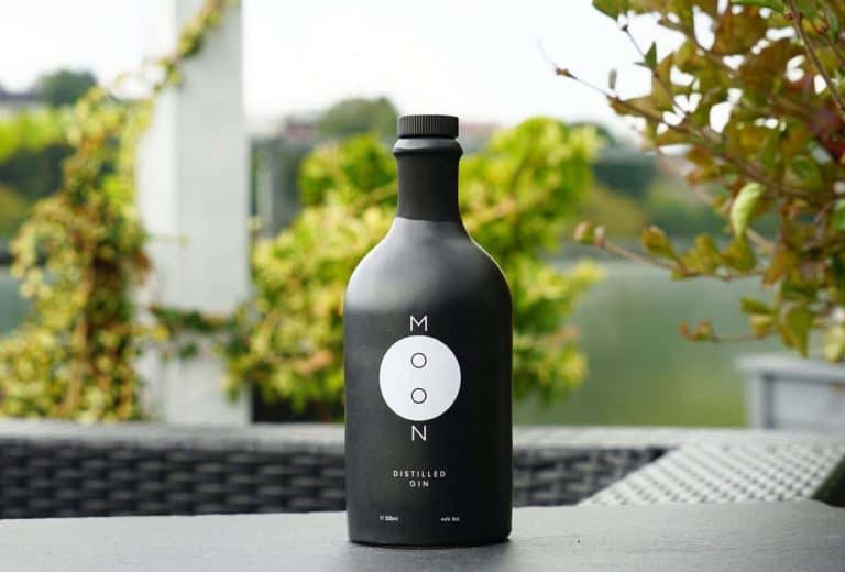 Auf einem Tisch im Freien steht eine schwarze Flasche mit der Aufschrift „MOON DISTILLED GIN“, im Hintergrund sind grüne Pflanzen und verschwommene Bäume zu sehen.
