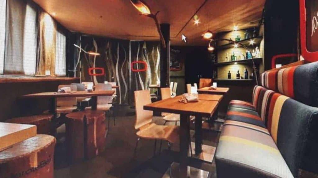 Ein gemütliches Restaurantinterieur mit Holztischen, gestreiften gepolsterten Bänken, modernen Stühlen, sanfter Beleuchtung und einer Rückwand, an der Flaschen in Regalen ausgestellt sind. Baumähnliche Dekorationen und große Fenster verleihen dem Raum eine natürliche Note.