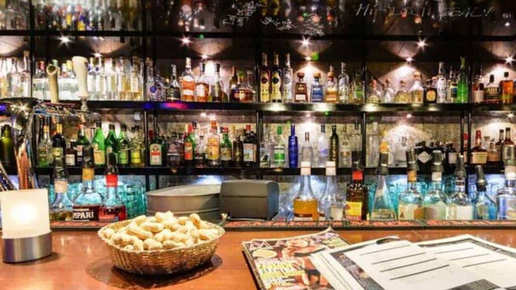 Eine gut beleuchtete Bar mit Regalen voller verschiedener Spirituosenflaschen hinter der Theke, einem Korb mit Snacks, einer Kerze, Zeitschriften und einer aufgeschlagenen Speisekarte auf der Theke.