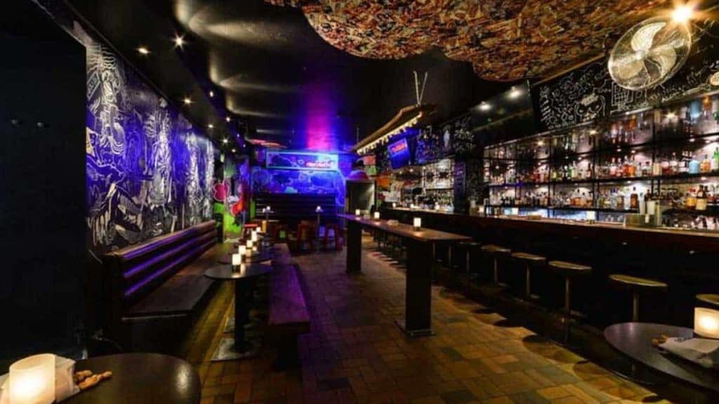 Eine schummrig beleuchtete Bar mit Barhockern an einer langen Theke, Kerzen auf den Tischen, kunstvollen Wandbildern und bunten Neonlichtern im hinteren Bereich, die eine gemütliche, künstlerische Atmosphäre schaffen.