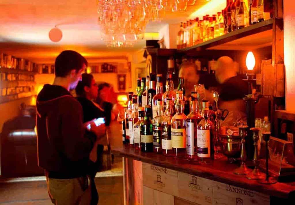 Eine schummrig beleuchtete Bar mit Spirituosenflaschen auf der Theke, Menschen, die stehen und sich unterhalten, und ein warmer orangefarbener Schein, der den Raum erfüllt. Über der Bar hängen Gläser.
