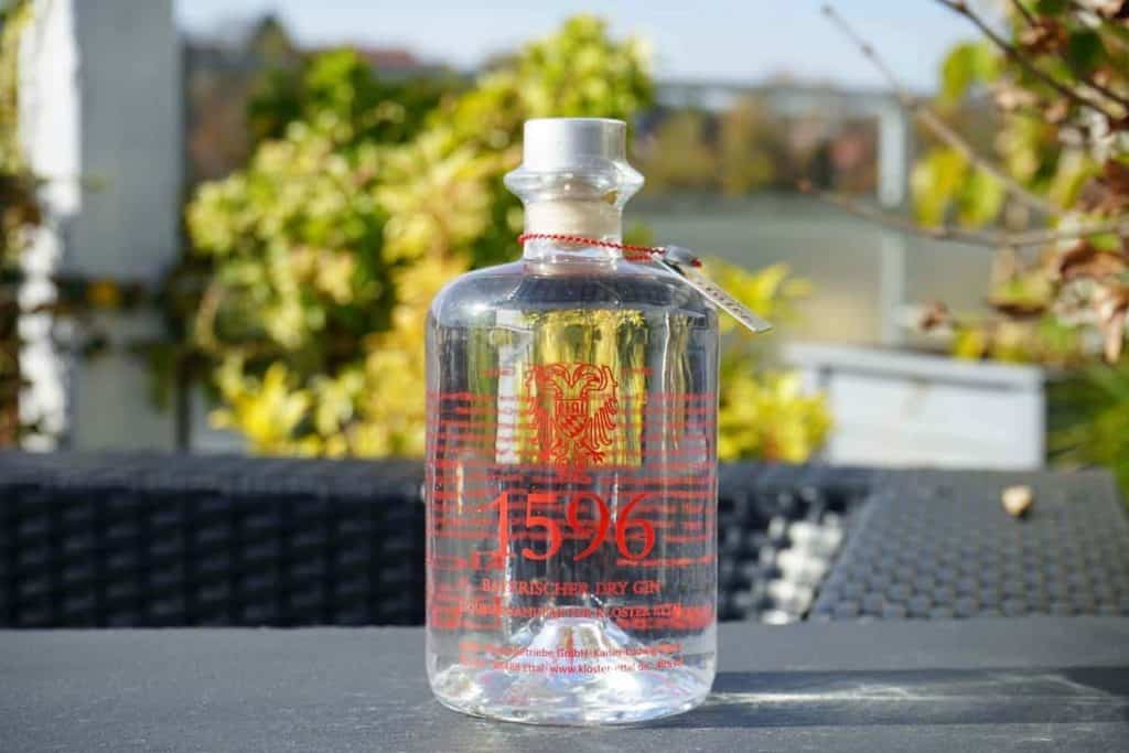 Auf einem Tisch im Freien steht eine klare Glasflasche mit der Aufschrift „1596 Saar Dry Gin“ in roter Schrift, im sonnigen Hintergrund sind verschwommene grüne Blätter und Gebäude zu sehen.