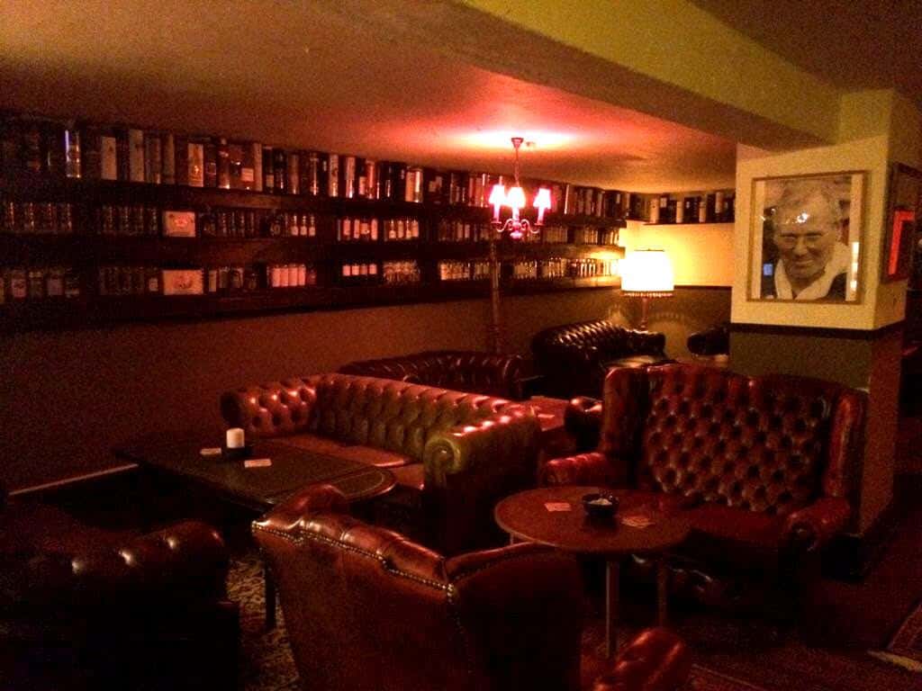 Ein schwach beleuchteter Wohnbereich mit braunen Ledersofas, Bücherregalen voller Bücher, einem Kronleuchter, einer Lampe und einem gerahmten Porträt an der Wand, die eine gemütliche, klassische Atmosphäre schaffen.