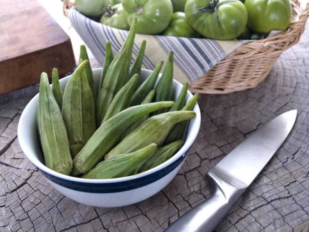 Auf einer Holzunterlage steht neben einem Messer eine weiße Schüssel, die mit frischen Okraschoten gefüllt ist. Im Hintergrund liegen in einem Korb mehrere grüne Tomaten.
