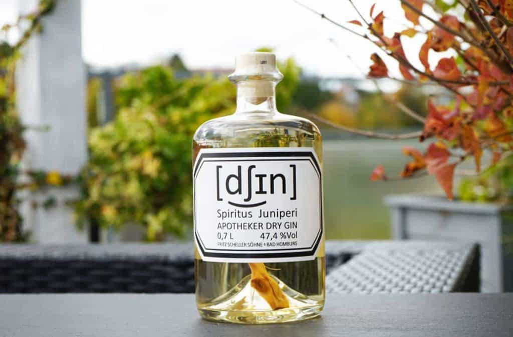 Auf einem Tisch im Freien steht eine Flasche „Apotheker“ Dry Gin mit der Aufschrift „Spiritus Juniperi“, im Hintergrund sind Herbstblätter und verschwommene Grünpflanzen zu sehen.