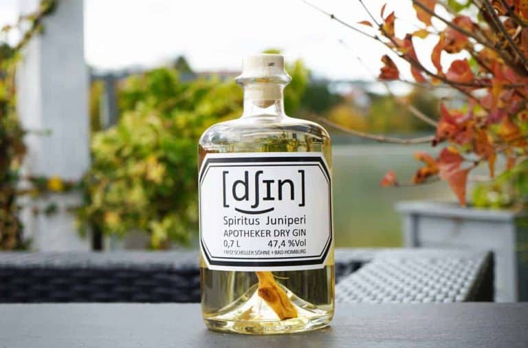 Auf einem Tisch im Freien steht eine Flasche „Apotheker“ Dry Gin mit der Aufschrift „Spiritus Juniperi“, im Hintergrund sind Herbstblätter und verschwommene Grünpflanzen zu sehen.