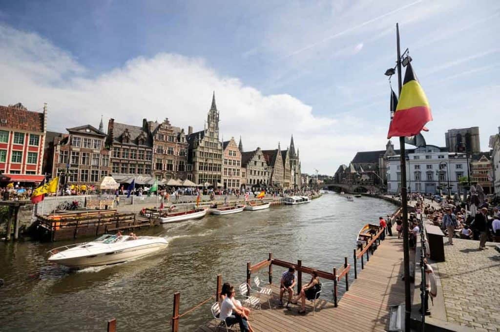 Eine lebhafte Szene am Flussufer einer europäischen Stadt mit historischen Gebäuden, Booten auf dem Wasser, Menschen, die am Kai entlangspazieren, und wehenden belgischen Flaggen an einem sonnigen Tag.
