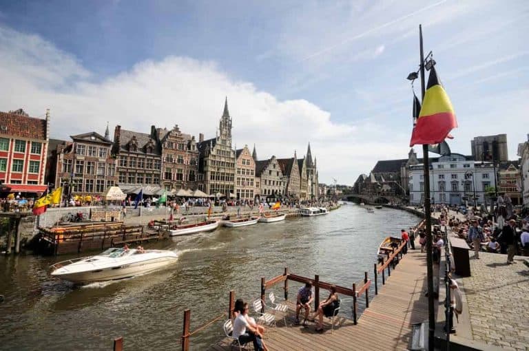 Eine lebhafte Szene am Flussufer einer europäischen Stadt mit historischen Gebäuden, Booten auf dem Wasser, Menschen, die am Kai entlangspazieren, und wehenden belgischen Flaggen an einem sonnigen Tag.
