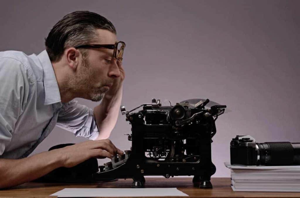 Ein Mann in einem hellen Hemd beugt sich vor und blickt durch seine Brille auf eine alte Schreibmaschine; neben ihm stehen auf dem Holztisch eine Kamera und Papierstapel vor einem schlichten grauen Hintergrund.