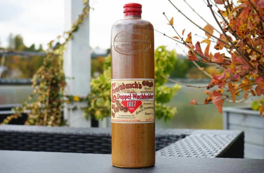 Auf einem schwarzen Tisch im Freien steht eine hohe braune Flasche mit der Aufschrift „Doppel-Wachholder 1817“; im Hintergrund sind Herbstblätter sowie ein verschwommener Hintergrund aus Grünpflanzen und einem Geländer zu sehen.