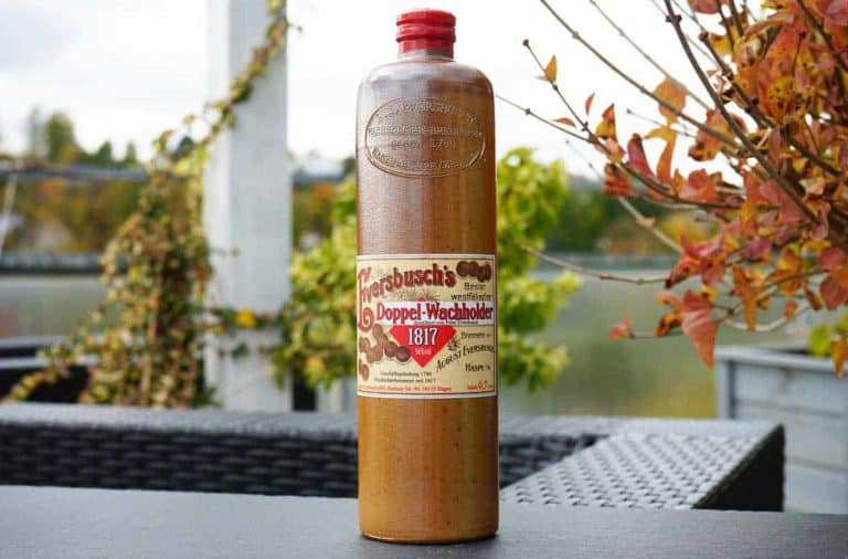 Auf einem schwarzen Tisch im Freien steht eine hohe braune Flasche mit der Aufschrift „Doppel-Wachholder 1817“; im Hintergrund sind Herbstblätter sowie ein verschwommener Hintergrund aus Grünpflanzen und einem Geländer zu sehen.