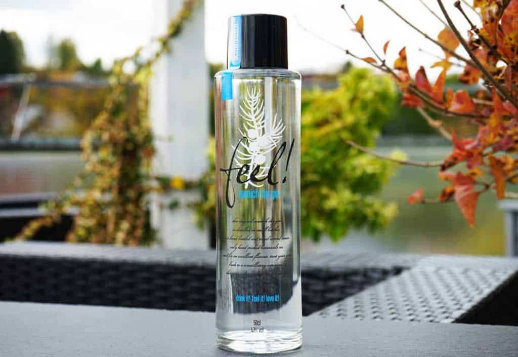 Auf einem Tisch im Freien steht eine durchsichtige Flasche „Feel!“-Mineralwasser, im Hintergrund sind grüne und orangefarbene Blätter zu sehen. Das Etikett zeigt ein weißes Blattmotiv und blauen Text.