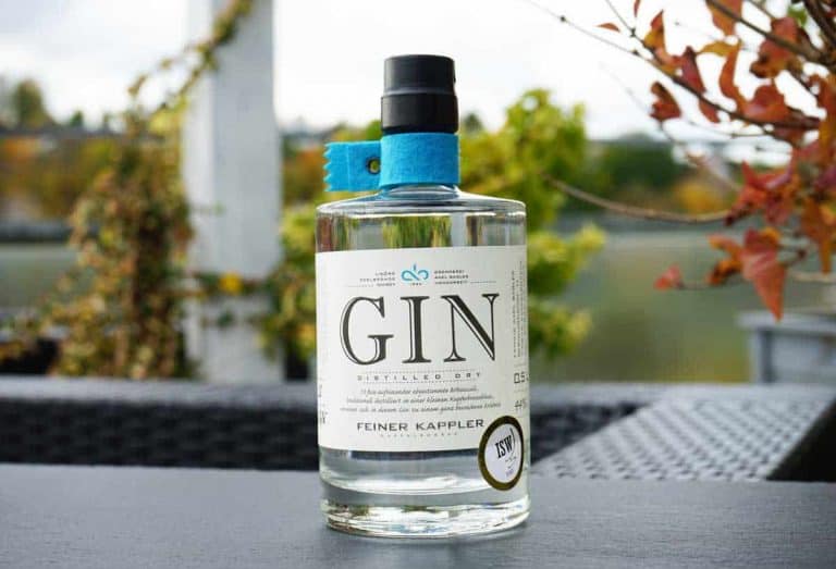 Auf einem Tisch im Freien steht eine durchsichtige Gin-Flasche mit der Aufschrift „GIN Distilled Dry Feiner Kappler“, im Hintergrund sind Grünpflanzen und verschwommene Blätter zu sehen.