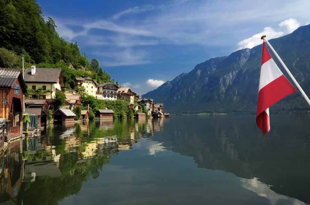 Ein Dorf am See mit bunten Häusern und Bergen im Hintergrund, die sich im ruhigen Wasser spiegeln. Im Vordergrund ist eine österreichische Flagge zu sehen.