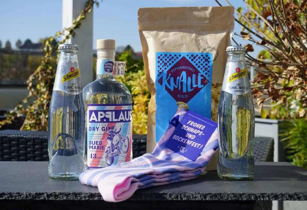 Zwei Flaschen Tonic Water, eine Flasche Gin, eine Tüte Snacks und ein Paar lila gestreifte Socken stehen auf einem Tisch im Freien, im Hintergrund sind Pflanzen und Stühle zu sehen.