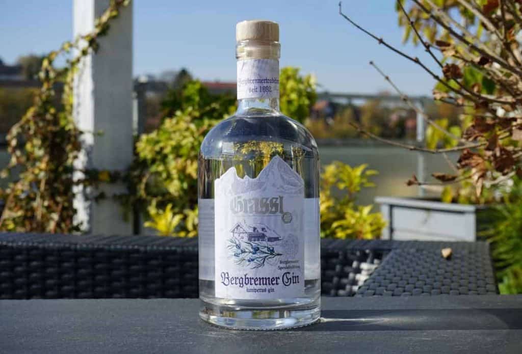 Auf einem dunklen Tisch im Freien steht eine durchsichtige Flasche Bergbrenner Gin, im unscharfen Hintergrund sind Pflanzen und ein Fluss zu sehen. Auf dem Etikett ist eine Illustration eines Berges und einer Berghütte abgebildet.