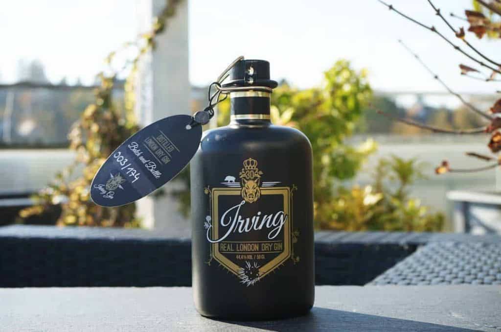 Eine schwarze Flasche „Spring Real London Dry Gin“ mit goldenem Etikett steht draußen auf einem Tisch. Am Flaschenhals ist ein Anhänger befestigt, und im Hintergrund sind verschwommene Pflanzen und ein Zaun zu sehen.