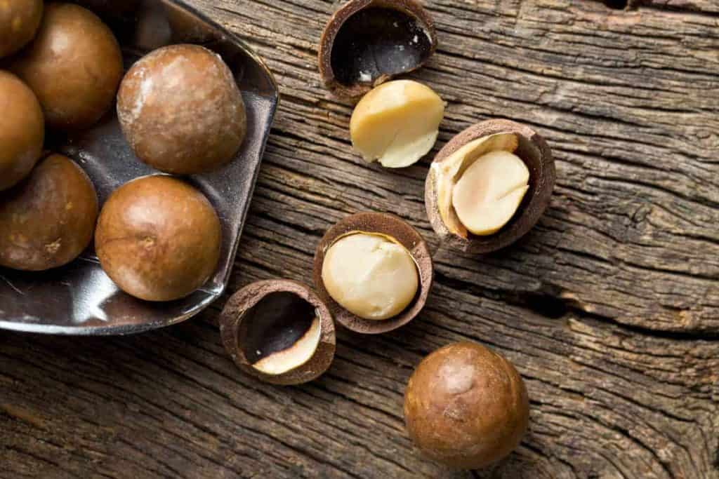Ganze und aufgebrochene Macadamia-Nüsse auf einer rustikalen Holzunterlage, wobei einige Nüsse ihr cremiges Inneres preisgeben, während andere noch in ihrer Schale stecken.
