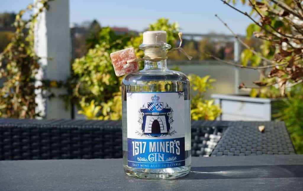 Auf einem Tisch im Freien steht eine durchsichtige Flasche „1517 Miners Gin“ mit einem Korken und einem kleinen Stein daran, umgeben von Grünpflanzen und unscharfen Hintergrundelementen.