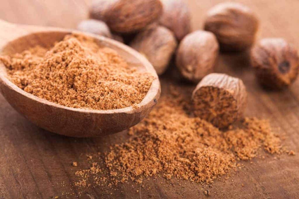 Ein Holzlöffel mit gemahlener Muskatnuss liegt auf einer Holzfläche, umgeben von ganzen Muskatnusskörnern.