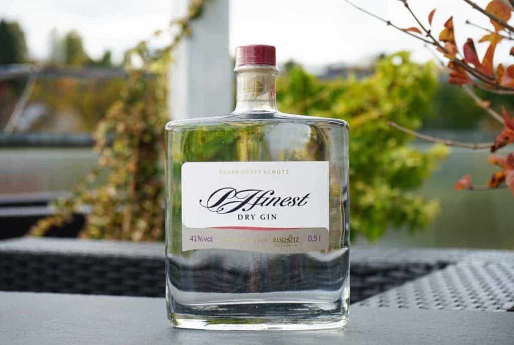 Auf einem Tisch im Freien steht eine Flasche „Afinest Dry Gin“ mit rotem Verschluss; im Hintergrund sind verschwommene grüne Pflanzen und ein Geländer zu sehen.