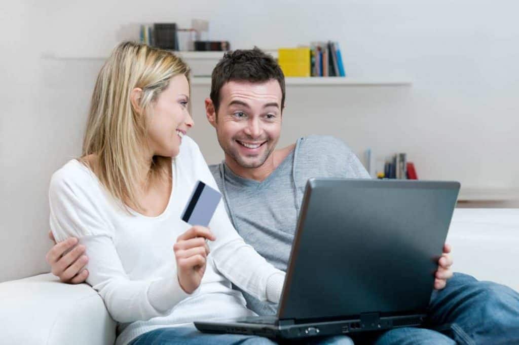Ein lächelndes Paar sitzt mit einem Laptop auf einem Sofa; die Frau hält eine Kreditkarte in der Hand, und der Mann schaut auf den Bildschirm, was darauf hindeutet, dass sie gemeinsam einkaufen oder einen Online-Kauf tätigen.