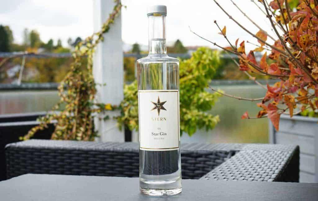 Auf einem schwarzen Gartentisch steht eine klare Glasflasche mit „Stjern Star Gin“, umgeben von Herbstlaub und Gartenmöbeln, im Hintergrund sind Grünpflanzen und ein Geländer zu sehen.