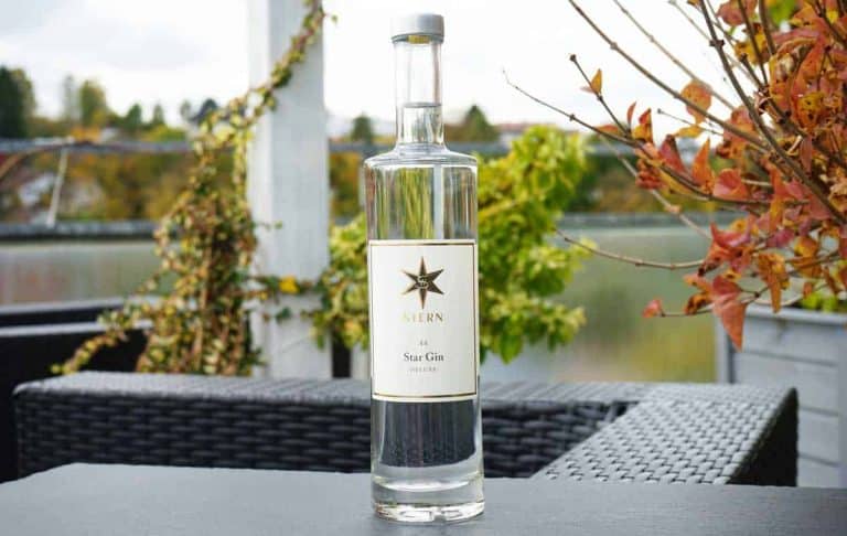 Auf einem schwarzen Gartentisch steht eine klare Glasflasche mit „Stjern Star Gin“, umgeben von Herbstlaub und Gartenmöbeln, im Hintergrund sind Grünpflanzen und ein Geländer zu sehen.