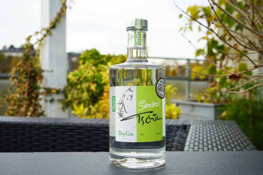 Auf einem schwarzen, geflochtenen Tisch im Freien steht eine durchsichtige Flasche „Stockers Dry Gin“ mit einem grün-weißen Etikett, umgeben von grünen Pflanzen und einem unscharfen Hintergrund.