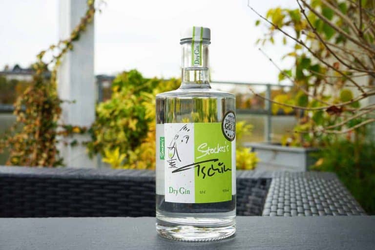 Auf einem schwarzen, geflochtenen Tisch im Freien steht eine durchsichtige Flasche „Stockers Dry Gin“ mit einem grün-weißen Etikett, umgeben von grünen Pflanzen und einem unscharfen Hintergrund.
