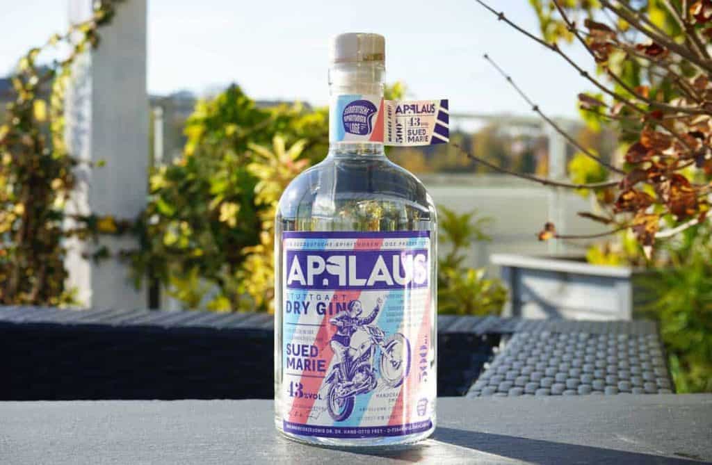 Auf einem Tisch im Freien steht eine Flasche „Applaus Dry Gin“ mit einem violett-blauen Etikett, umgeben von Grünpflanzen und einem verschwommenen Hintergrund im Freien.